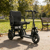 Black electric wheelchair on a paved path in a park with trees and a bench in the background.