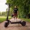 Man walking with a helmet and Teverun Fighter 11 electric scooter on a path in a park.