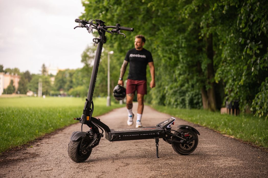 Man walking with a helmet and Teverun Fighter 11 electric scooter on a path in a park.