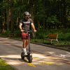 Person riding a Teverun Fighter 11 scooter on a paved path in a park with trees and a bench in the background.