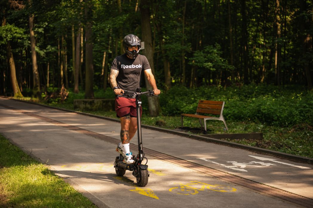 Person riding a Teverun Fighter 11 scooter on a paved path in a park with trees and a bench in the background.