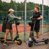 Two people with electric scooters on a paved area near a fence and trees.