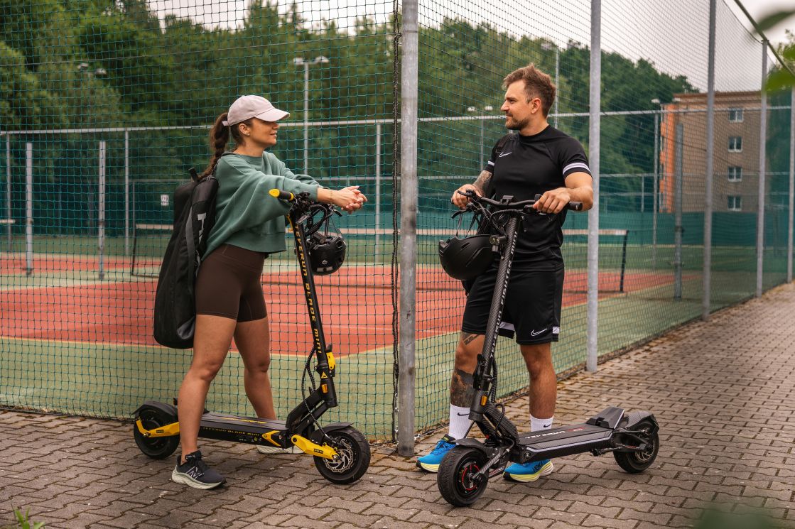Two people with electric scooters on a paved area near a fence and trees.