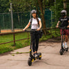 Two people riding Teverun electric scooters on a path with trees and a fence in the background.