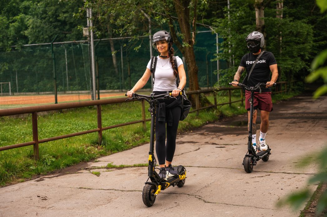 Two people riding Teverun electric scooters on a path with trees and a fence in the background.