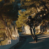Person riding a Teverun Fighter Supreme Ultra electric scooter on a mountain road with trees and rocks.