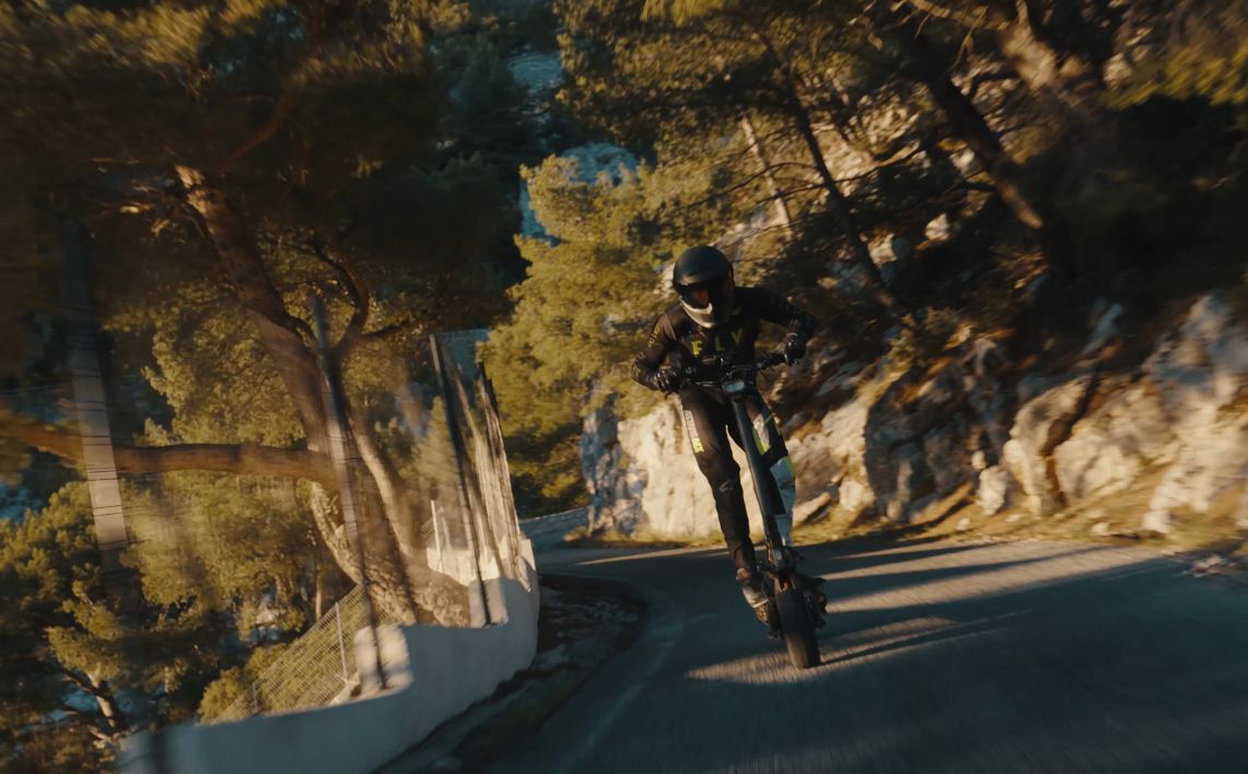 Person riding a Teverun Fighter Supreme Ultra electric scooter on a mountain road with trees and rocks.